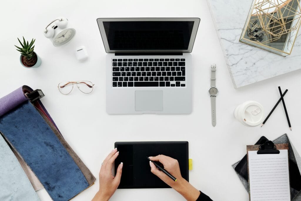 Top view of a workspace with a laptop, drawing tablet, and various desk items on a white surface.